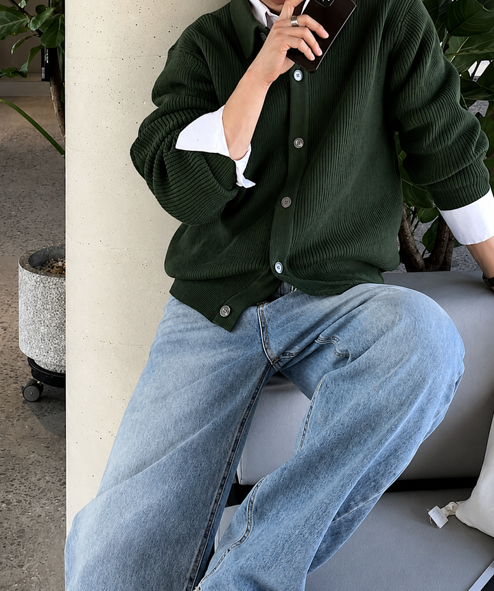 グリーンの襟付きカーディガンを着用した男性が、スマートフォンを片手に持ちながら座っている、リラックスしたサイジングが伝わるミドルショット。