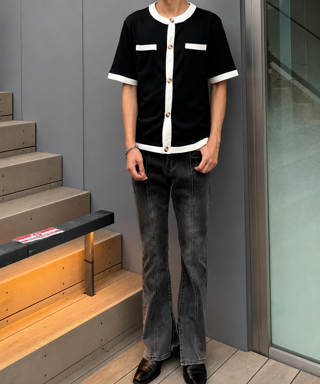 センターシームと裾スリットが特徴のブラックフレアデニムパンツを着用した男性の全身正面。白の配色が特徴的な黒の半袖シャツを着用。屋外のウッドデッキと階段前。