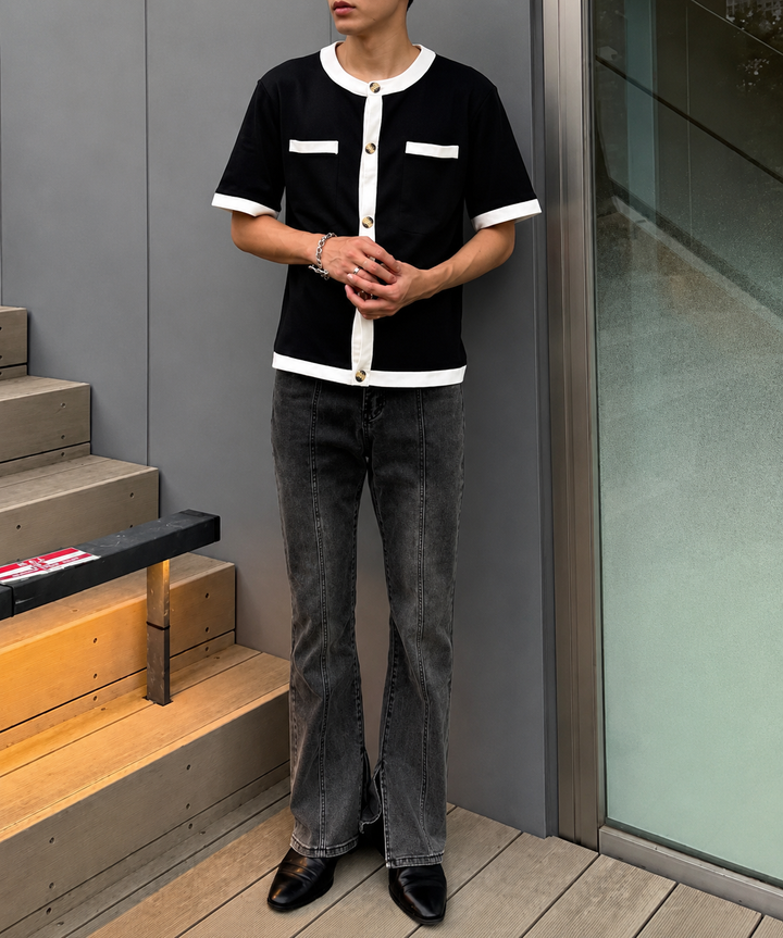 センターシームと裾スリットが特徴のブラックフレアデニムパンツを着用した男性の全身正面。白の配色が特徴的な黒の半袖シャツを着用。屋外のウッドデッキと階段前。ブレスレットとリングをコーディネート。