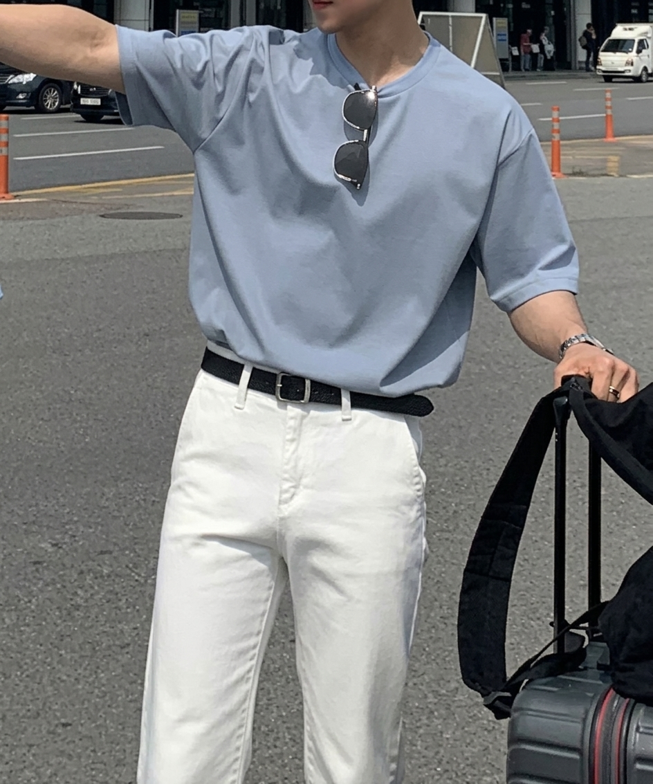 ライトブルーのクルーネックTシャツにサングラスを掛け、ホワイトのパンツと黒のベルトを合わせてスーツケースを持つ男性の正面。