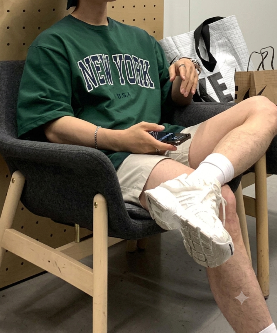 グレーの椅子に腰掛け、ダークグリーンのロゴTシャツと白スニーカーを着用してスマホを触る男性のカット