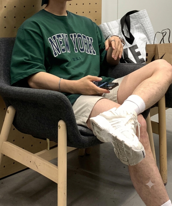 グレーの椅子に腰掛け、ダークグリーンのロゴTシャツと白スニーカーを着用してスマホを触る男性のカット