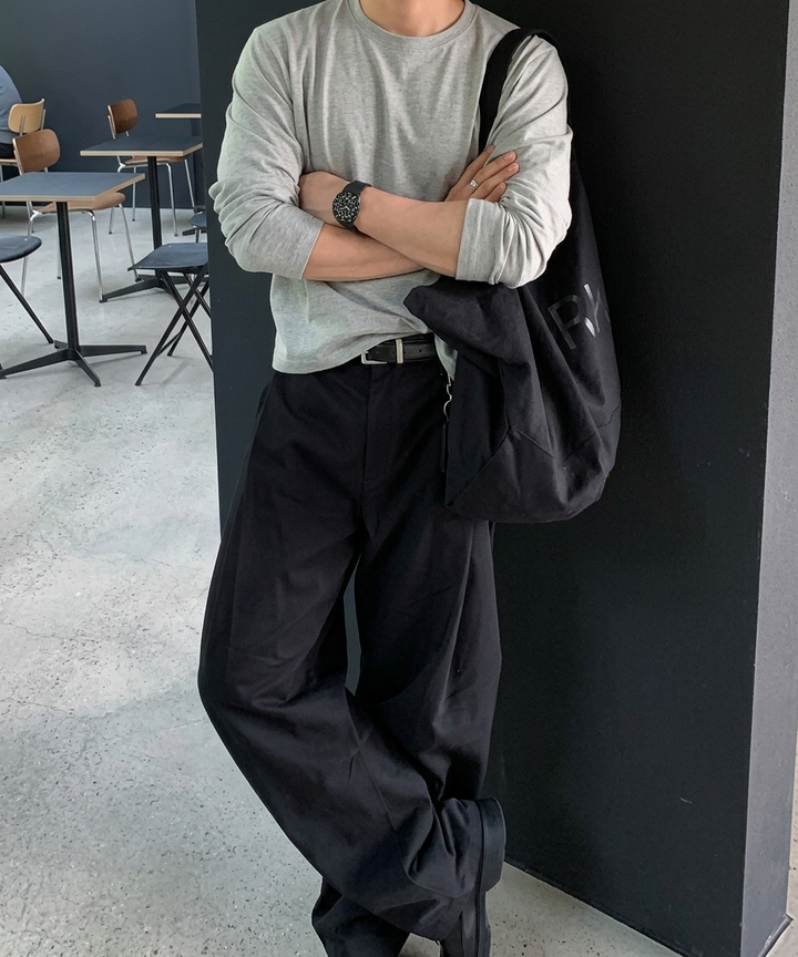 黒い壁に寄りかかり、腕を組んで足を交差させる男性の全身コーディネート。グレー、ブラック、レザーの小物が調和した、モードでアーバンなストリートスタイル