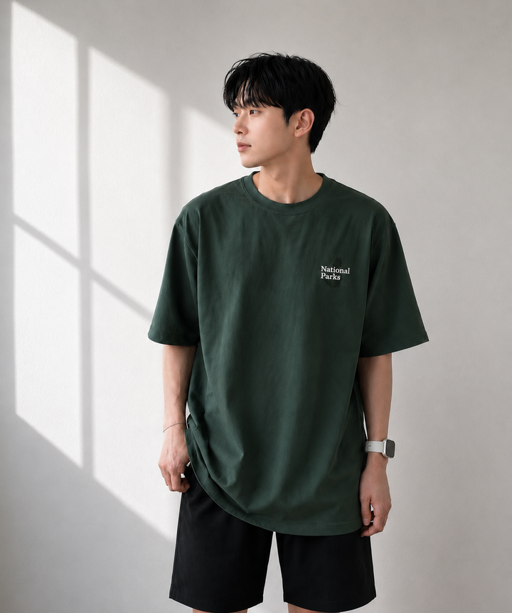 室内でダークグリーンのグラフィックTシャツの製品を着用した男性の半身ショット。ドロップショルダーのゆったりとしたシルエットと、左胸に施されたホワイトの「National Parks」ミニロゴデザインがクリアに伝わるフロントビュー。
