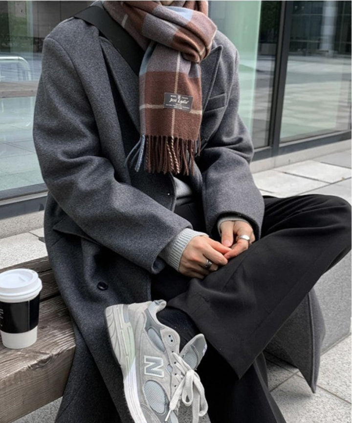 ダークグレーのロングチェスターコートに黒のパンツ、グレーのスニーカーを合わせ、手元にコーヒーカップを置いた男性のアップカット。