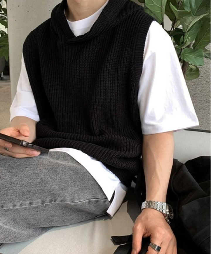 白Tシャツとブラックのフード付きベストをレイヤードし、グレーのパンツを穿いてベンチに腰掛け、左手でスマートフォンを持ちながら過ごす男性のアップ