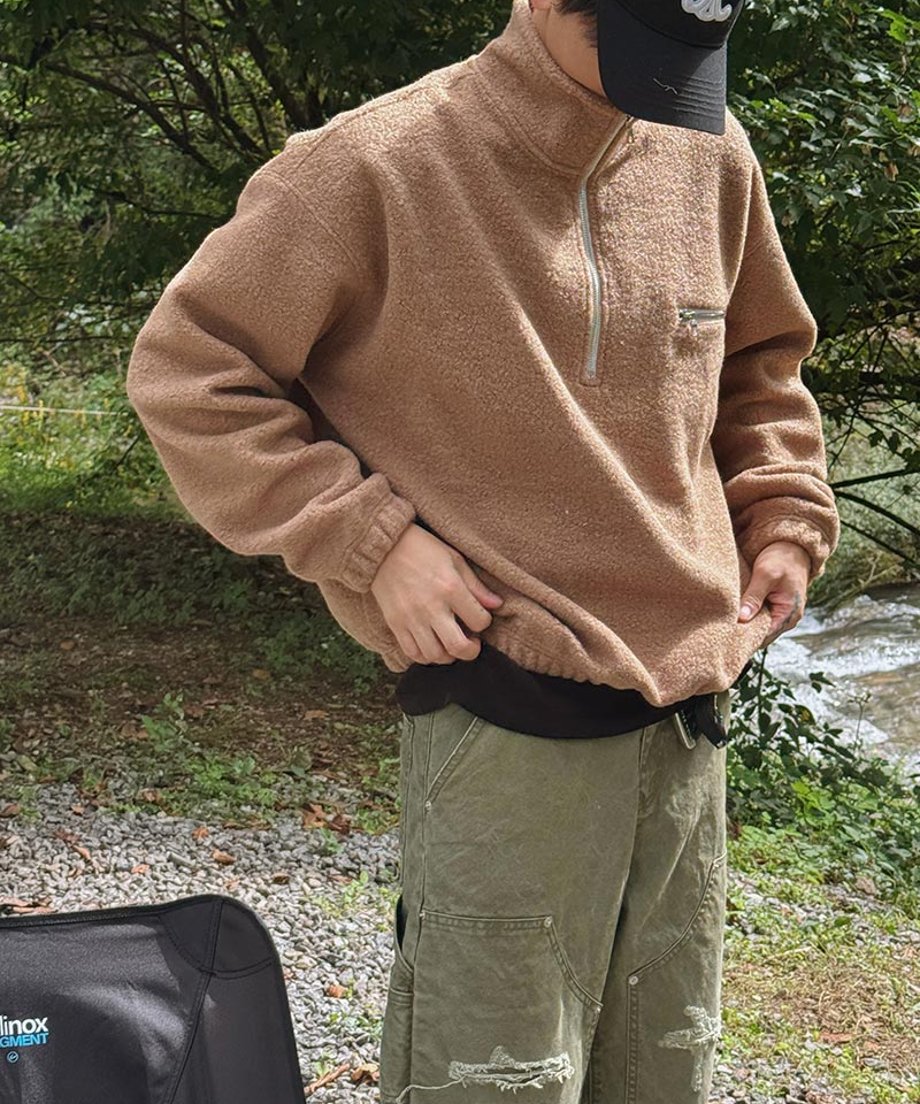 ベージュのハーフジップフリースに黒のキャップを被り、カーキのカーゴパンツのウエスト周りを整えている男性の、川沿いでのスタイリングカット。