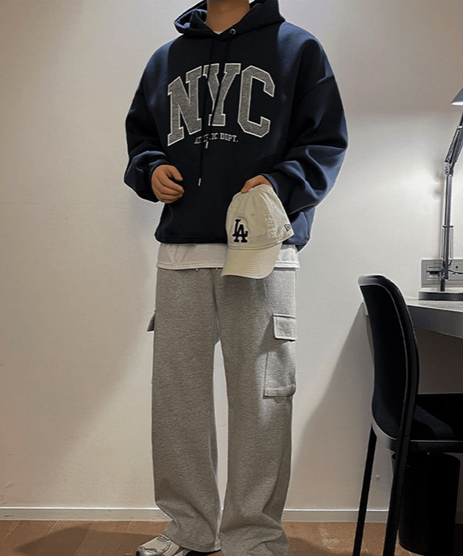 グレーのスウェットパンツのポケットに手を入れ、ネイビーブルーのNYCロゴパーカーをラフに着こなす男性のフロントカット。