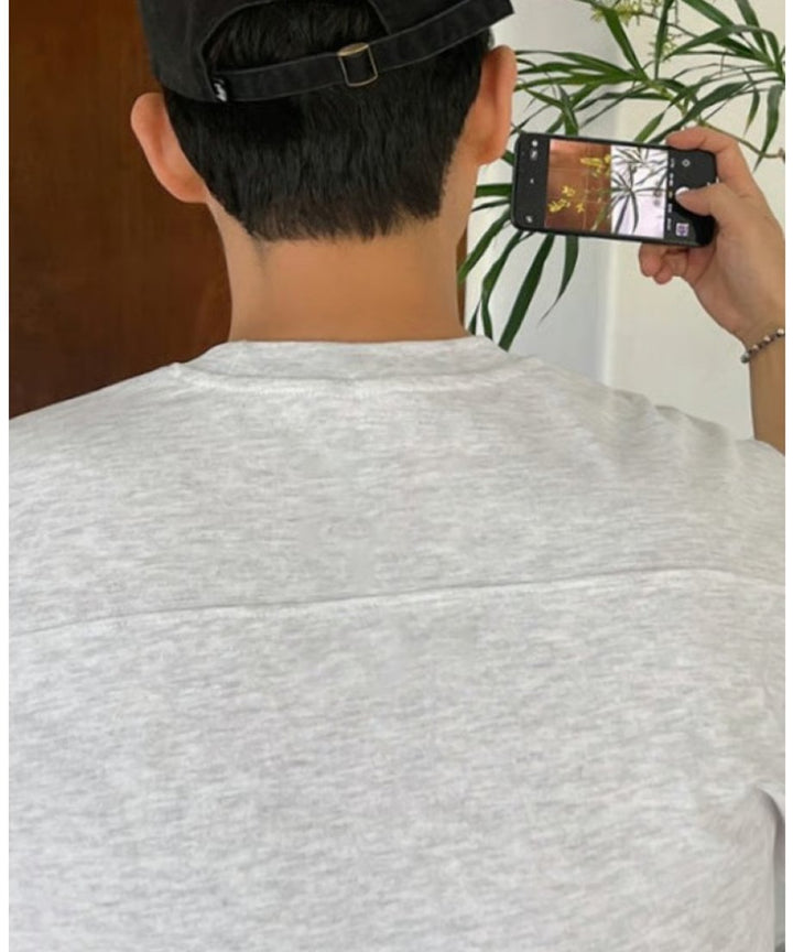 フラワーグレーのTシャツに黒のキャップを後ろ被りした男性が、スマートフォンで前方の植物を撮影している様子のバックビュー（後頭部から肩口のアップ）。