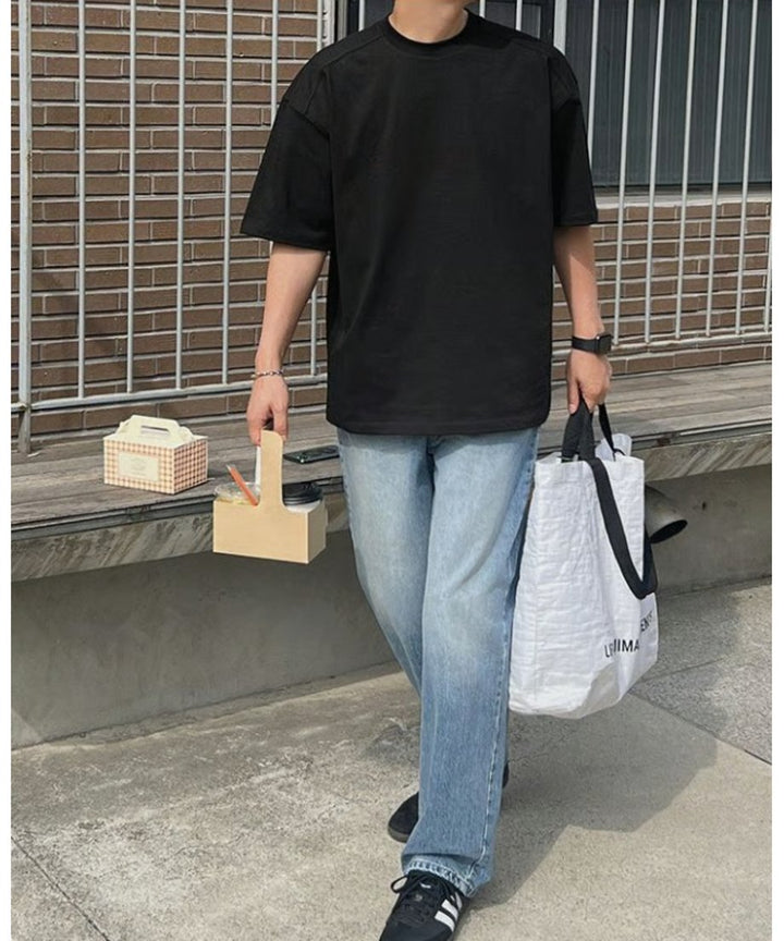 ブラックのルーズフィットTシャツにライトインディゴのワイドデニムパンツ、黒のスニーカーを合わせ、左手に白いトートバッグ、右手にドリンクホルダーを持って歩く男性の正面スタイリング。