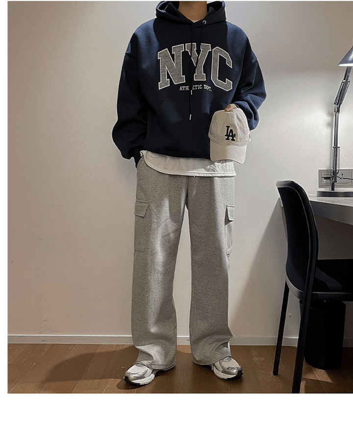 ネイビーブルーのNYCロゴパーカーに、白Tシャツをレイヤードし、グレーのカーゴスウェットパンツを合わせたメンズストリートスタイル。手に持ったベージュのLAキャップがアクセント。