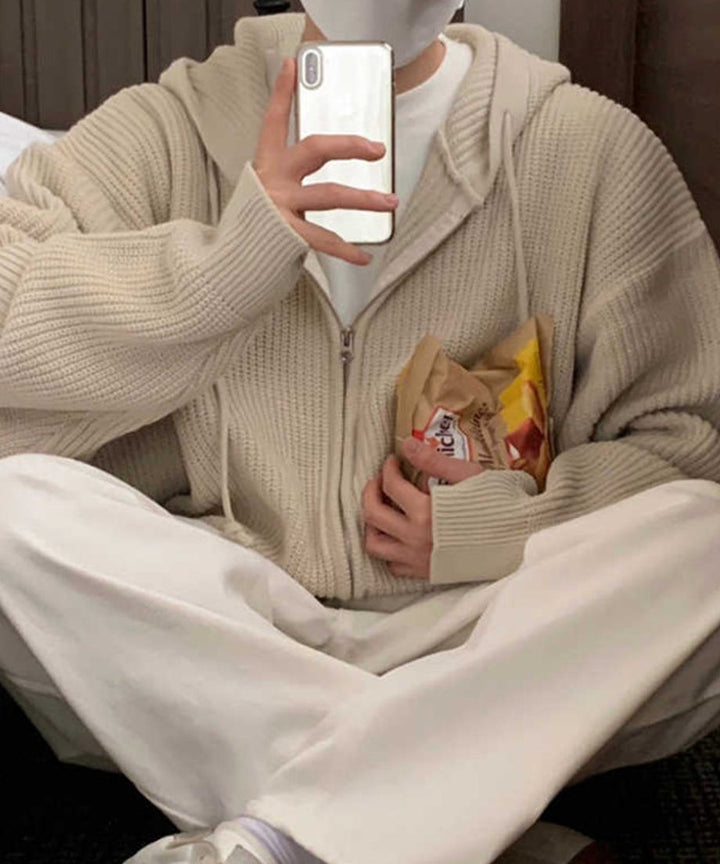 ベージュのニットパーカーのポケット付近にお菓子の袋を持ちながらスマートフォンを構える男性のリラックスしたカット。