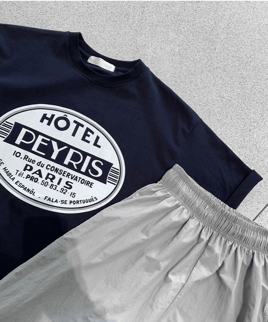 スタイリッシュなコンクリート調の床に平置きされた、ネイビーのサークルロゴTシャツとライトグレーのナイロン風イージーパンツ。