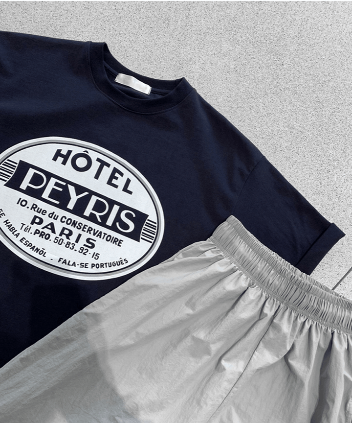 スタイリッシュなコンクリート調の床に平置きされた、ネイビーのサークルロゴTシャツとライトグレーのナイロン風イージーパンツ。