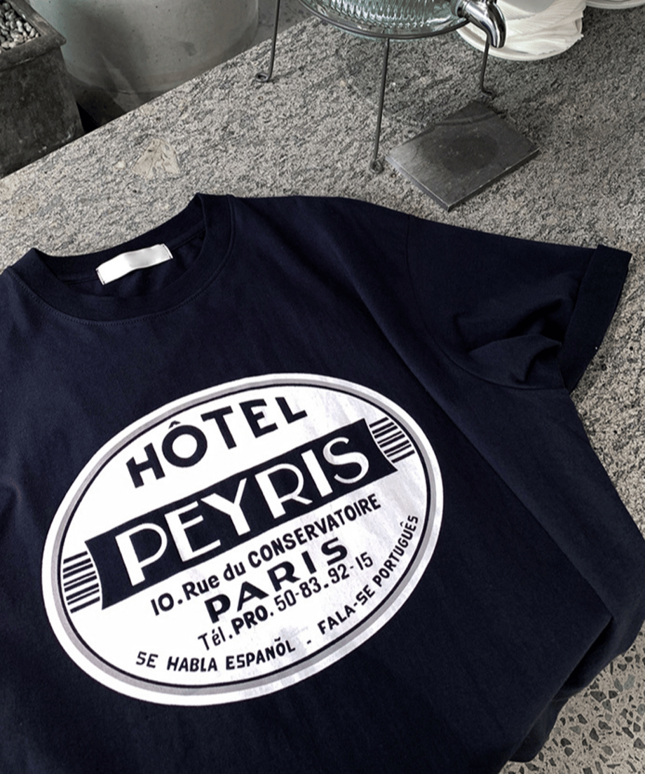 石目調のカフェカウンターのような床に平置きされた、ネイビーのサークルロゴTシャツ。フロントに大きくプリントされた「HÔTEL PEYRIS PARIS」の白いサークルロゴのディテールがはっきりと分かる、質感重視の接写カット。