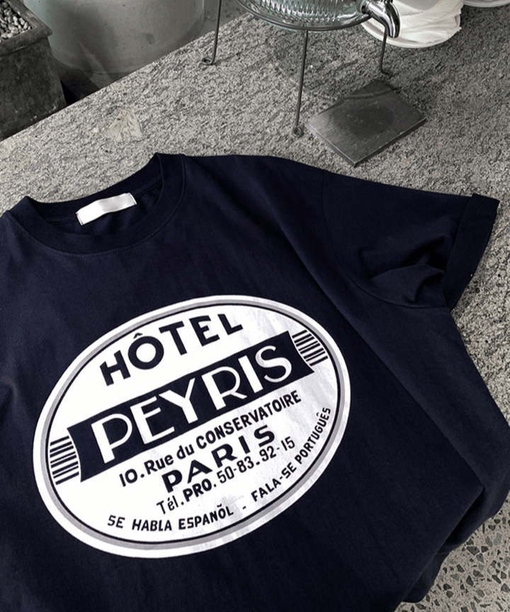 石目調のカフェカウンターのような床に平置きされた、ネイビーのサークルロゴTシャツ。フロントに大きくプリントされた「HÔTEL PEYRIS PARIS」の白いサークルロゴのディテールがはっきりと分かる、質感重視の接写カット。