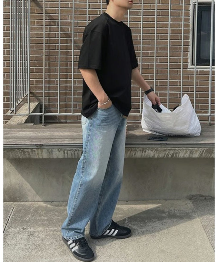 ブラックのルーズTシャツにライトインディゴのワイドデニム、黒のスニーカーを合わせ、ポケットに手を入れながら白いトートバッグの横に立つ男性のサイドビュー。