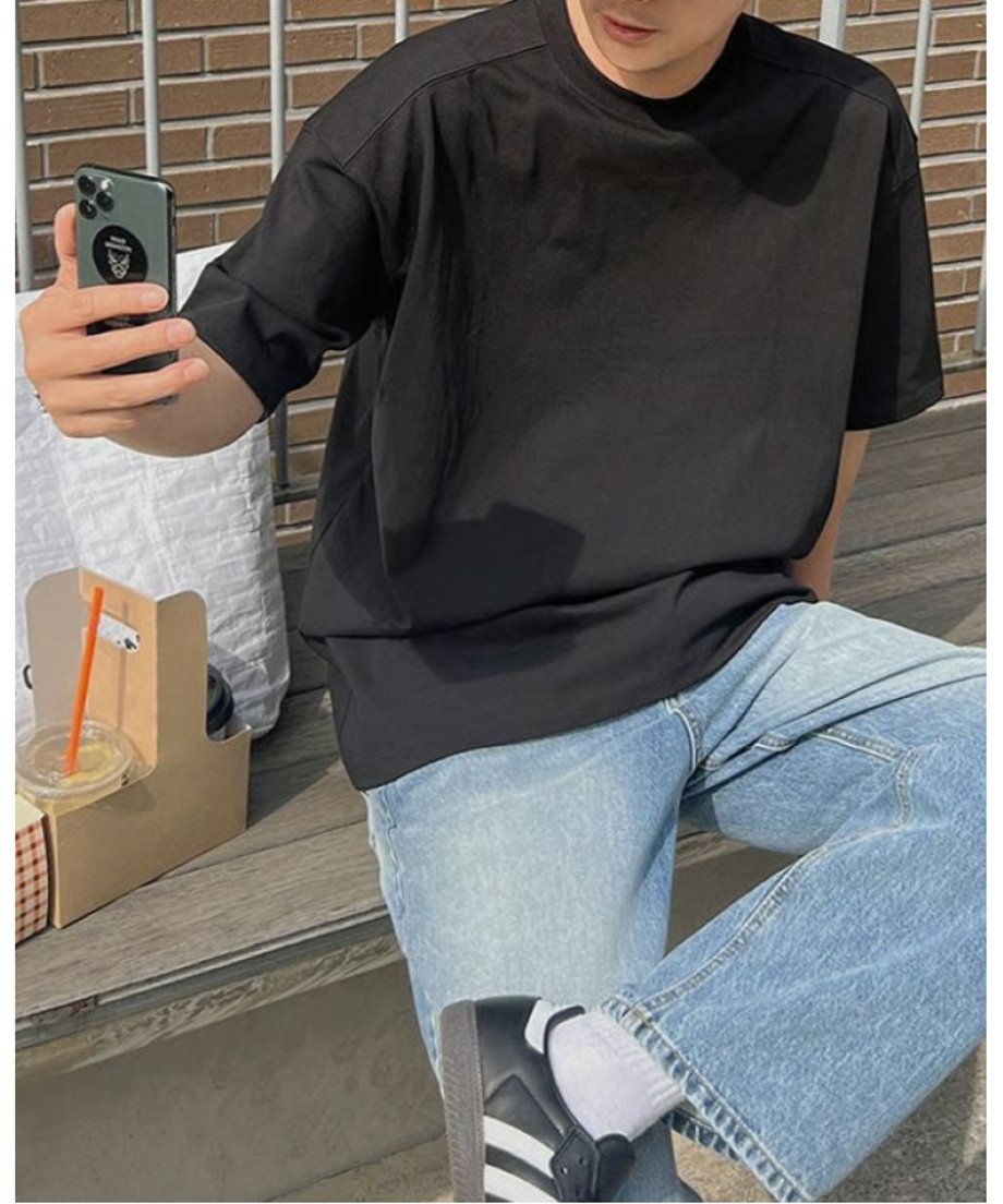 ブラックの半袖Tシャツにライトインディゴのデニムパンツ、黒のスニーカーを合わせ、ベンチに腰掛けながら右手でスマートフォンを持って自撮りをする男性の斜めからのアップ。