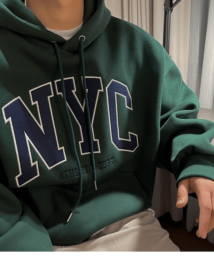 ダークグリーンのパーカーの胸元に施された、ネイビーに白の縁取りがある「NYC」のビッグロゴワッペンと、その下の「ATHLETIC DEPT.」の刺繍ディテールを強調した接写カット。