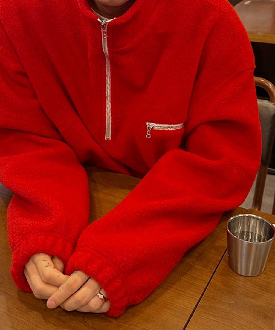 レッドのハーフジップフリースを着用し、木目調のテーブルに両腕を置いて座っている男性の手元のアップカット。