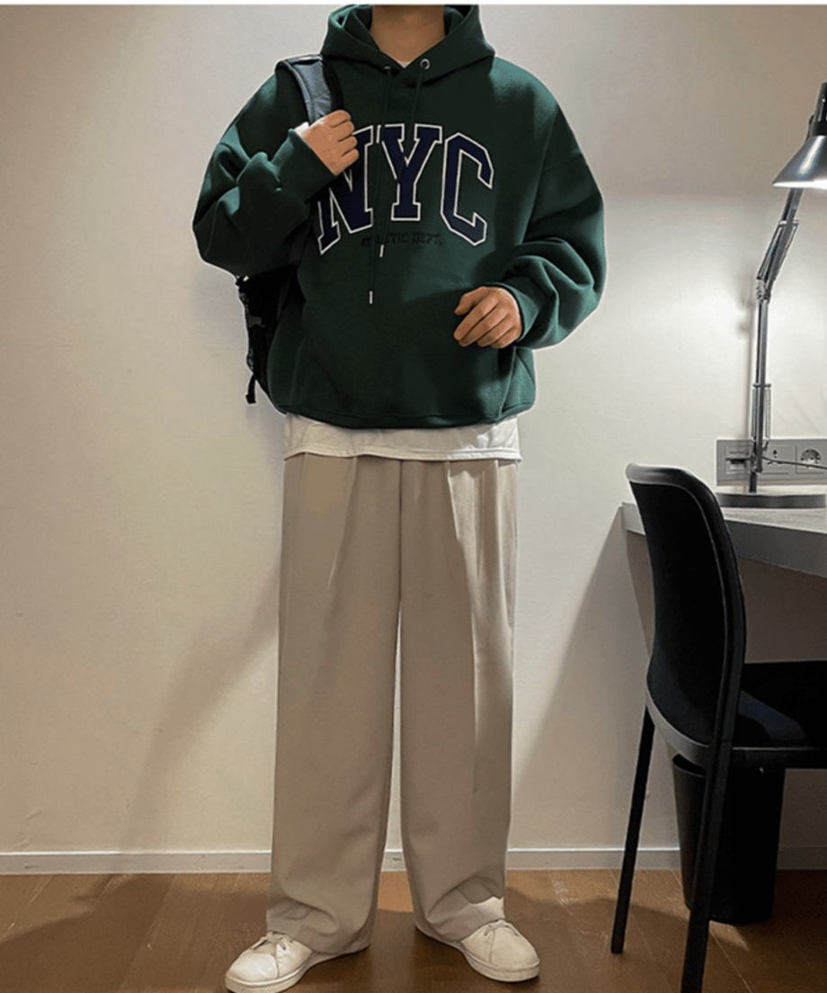 ダークグリーンのNYCロゴパーカーに白のインナーをレイヤードし、ベージュのタックワイドパンツ、白スニーカー、黒のリュックを合わせた男性の全身立ち姿カット。