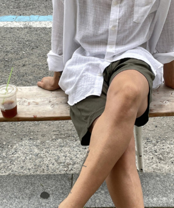 木製のベンチに腰掛け、白シャツにアーミーグリーンのショートパンツを合わせて足を組んでいる男性の足元のアップ