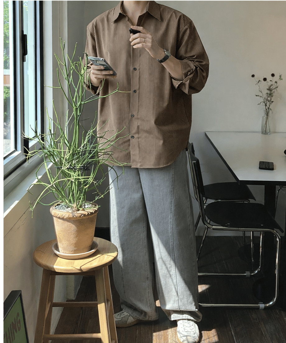 ブラウンのストライプ柄長袖シャツにグレーのワイドパンツを合わせ、室内の観葉植物の横でスマートフォンを手にする男性の全身スタイリング。