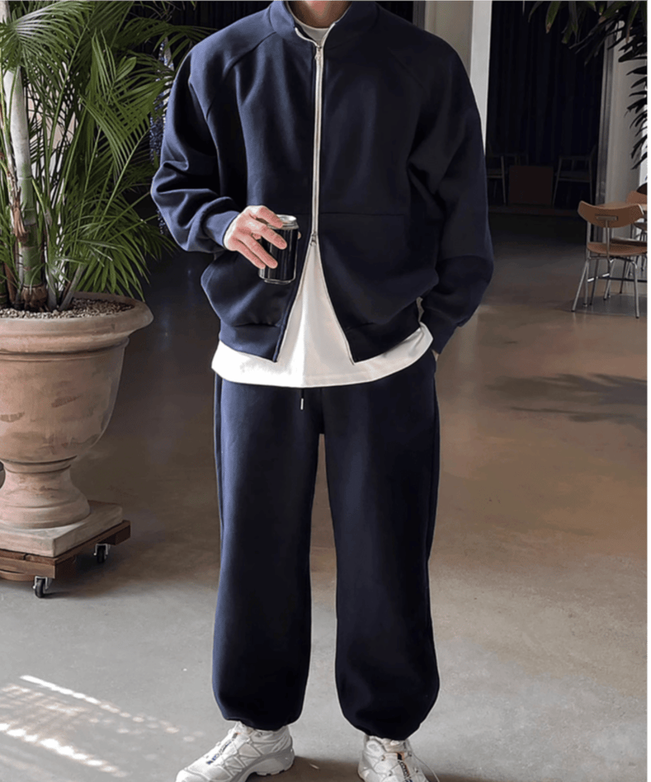 室内でネイビーのジップアップジャケットとトラックパンツのセットアップを着用し、右手に缶を持って立つ男性の全身スタイリング