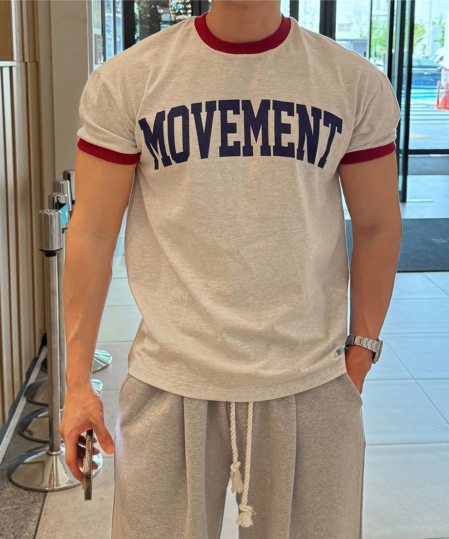 オートミール地に赤のリブ、ネイビーのロゴが入ったリンガーTシャツを着用し、グレーのスウェットパンツのポケットに左手を入れた男性の正面アップカット。