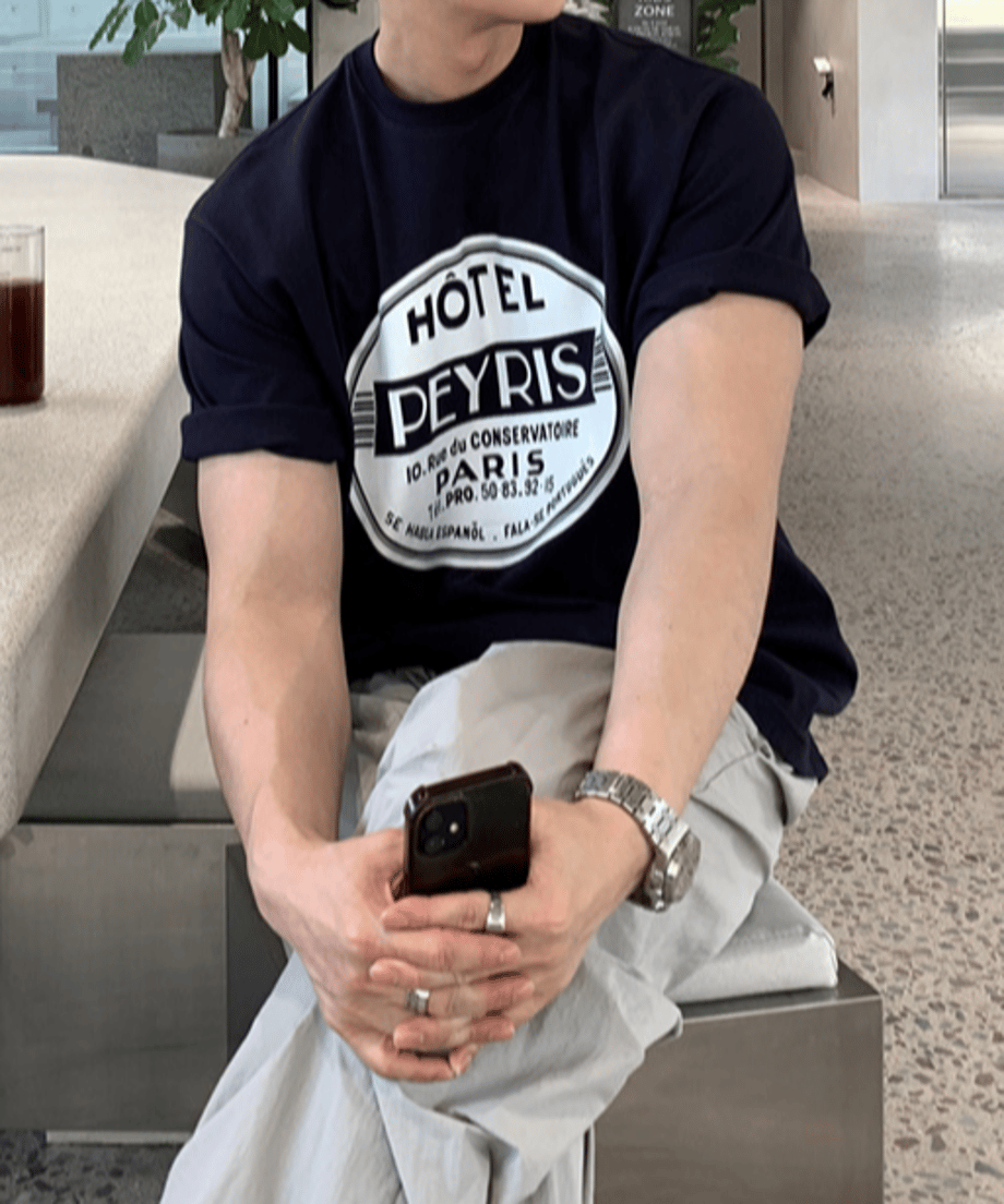 ネイビーのレトロホテル風サークルロゴTシャツに、ライトグレーのワイドイージーパンツを合わせた男性が、カフェの椅子に座ってスマートフォンを手に持っている手元のアップカット。