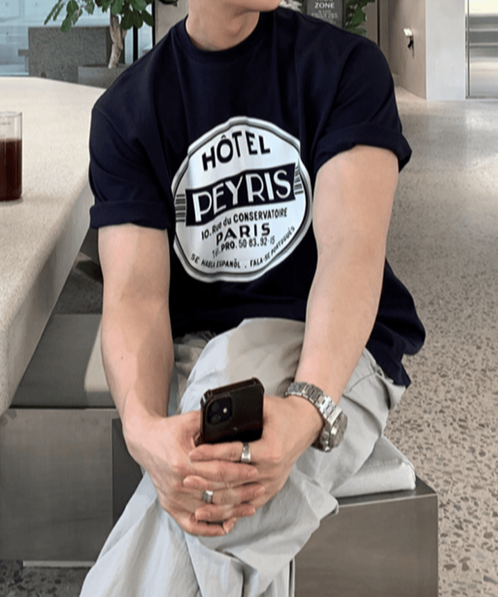 ネイビーのレトロホテル風サークルロゴTシャツに、ライトグレーのワイドイージーパンツを合わせた男性が、カフェの椅子に座ってスマートフォンを手に持っている手元のアップカット。