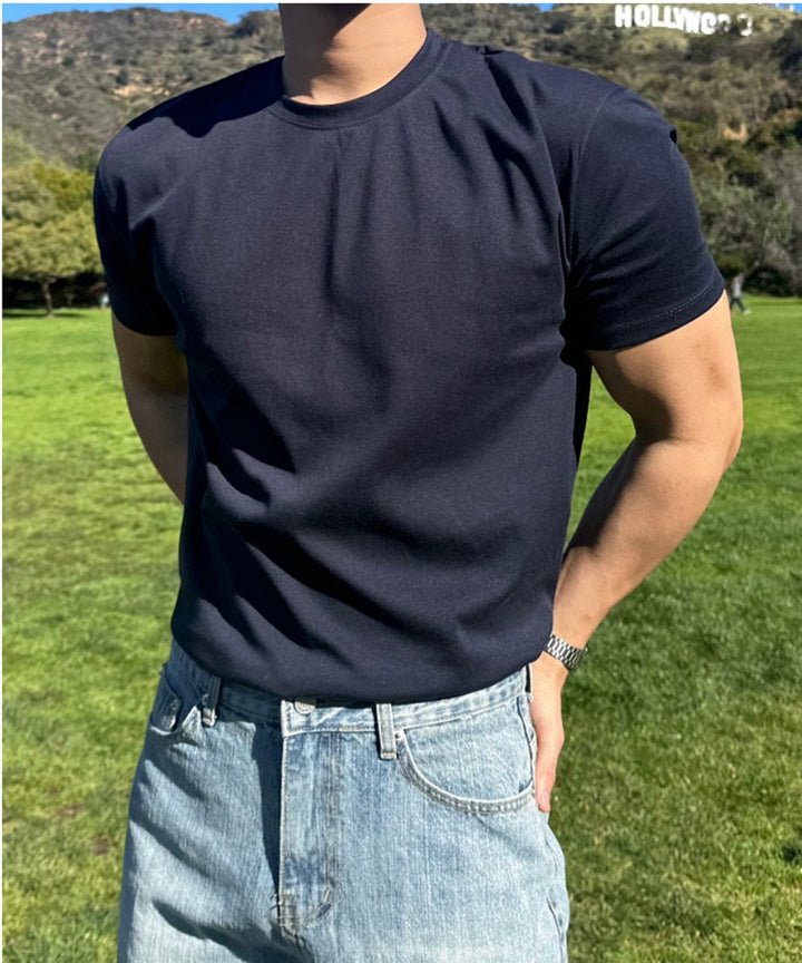 ネイビーの半袖クルーネックTシャツをライトブルーのデニムパンツにタックインし、緑の芝生をバックに立つ男性の正面からの上半身ショット。