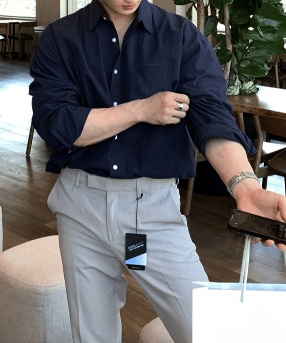 ネイビーの長袖シャツの袖をたくし上げ、右手にスマートフォン、左手に白いショップバッグを持って立っている男性のフロントアップカット。