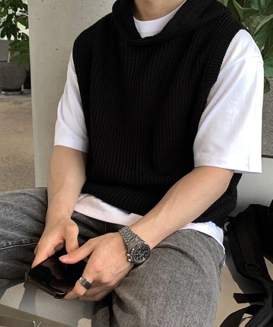 白Tシャツにブラックのフード付きニットベストをレイヤードし、グレーのパンツを穿いてベンチに座りながら両手でスマートフォンを持つ男性の手元のアップ