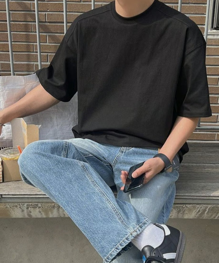 ブラックのルーズフィットTシャツにライトインディゴのデニムパンツ、黒のスニーカーを合わせ、ウッドデッキに腰掛けてスマホを持つ男性の正面スタイリング。