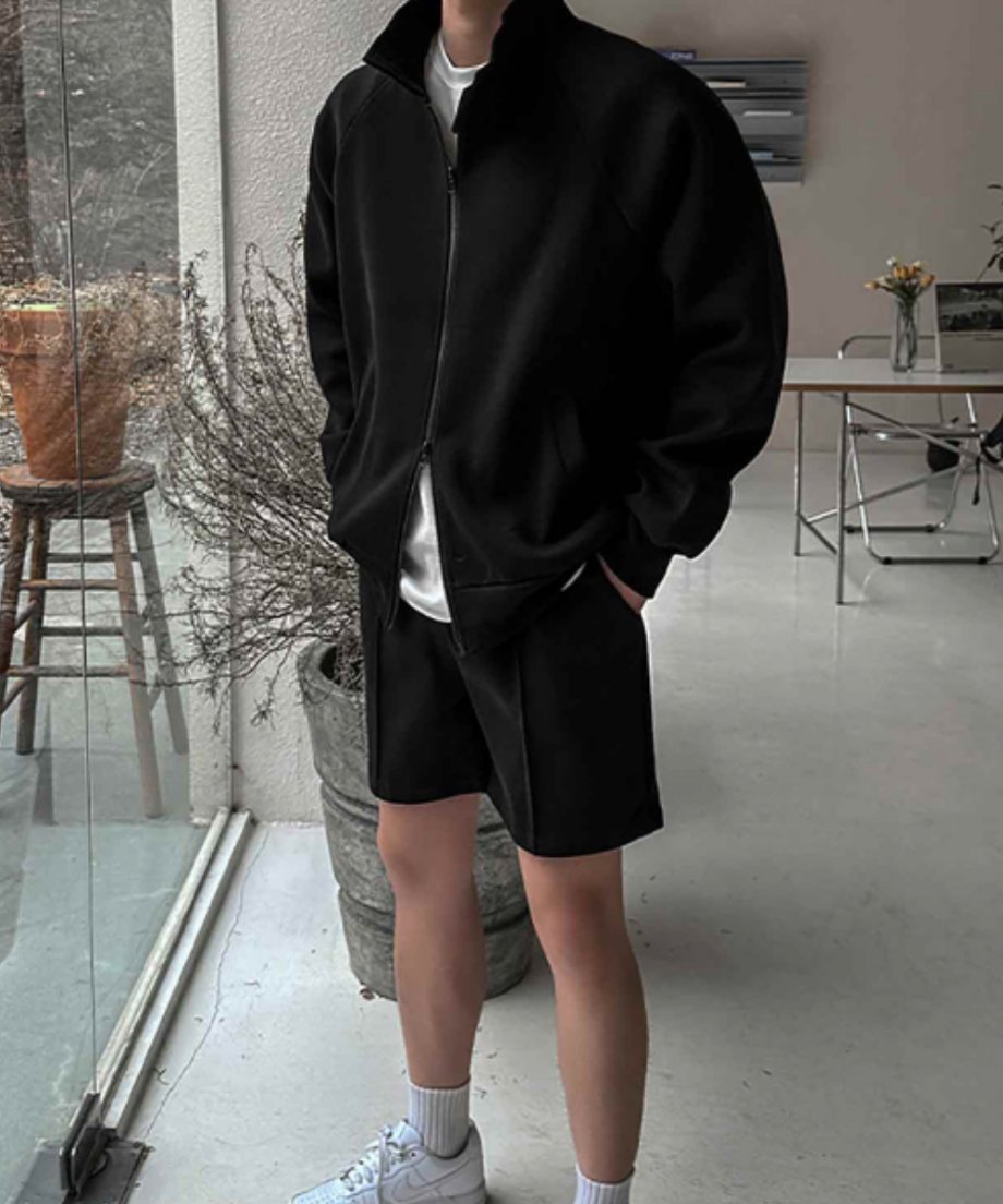 屋内で黒のスタンドカラージップアップジャケットと黒のショートパンツのセットアップを着用した男性のフロントスタイル