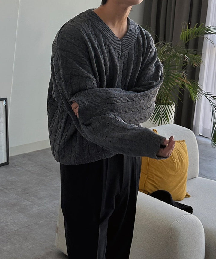 ケーブル編みのダークグレーVネックニットを着用し、腕組みをしながら白いソファの前に佇む男性のアップカット。