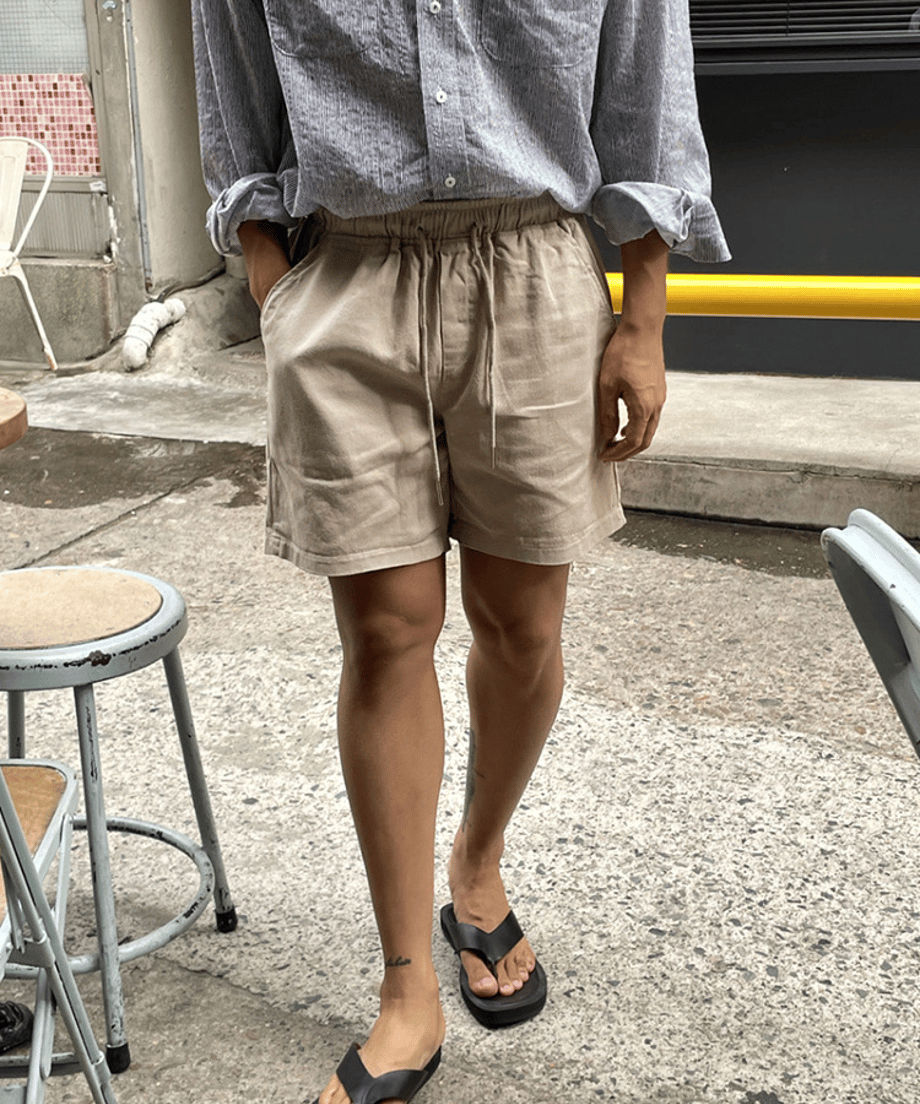 屋外でストライプシャツにベージュのショートパンツ、黒のトングサンダルを着用した男性のフロントスタイル