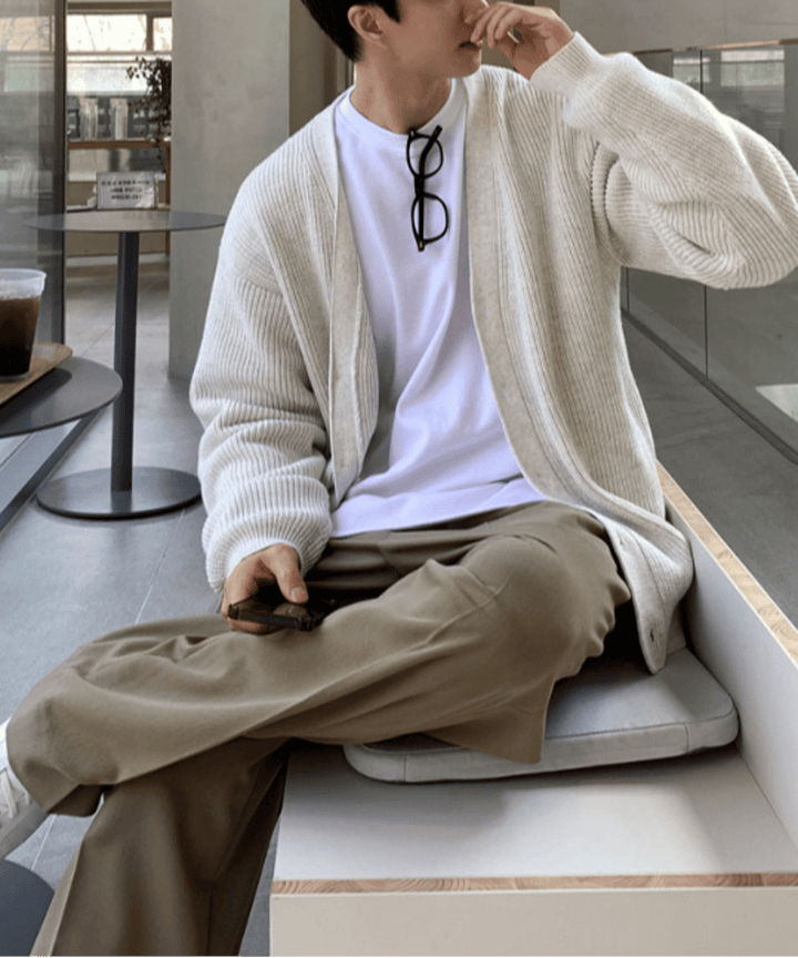 胸元にメガネを掛けた白いTシャツにカーディガンを羽織り、ダークベージュのワイドパンツを穿いて足を組んで座る男性の正面アップ。