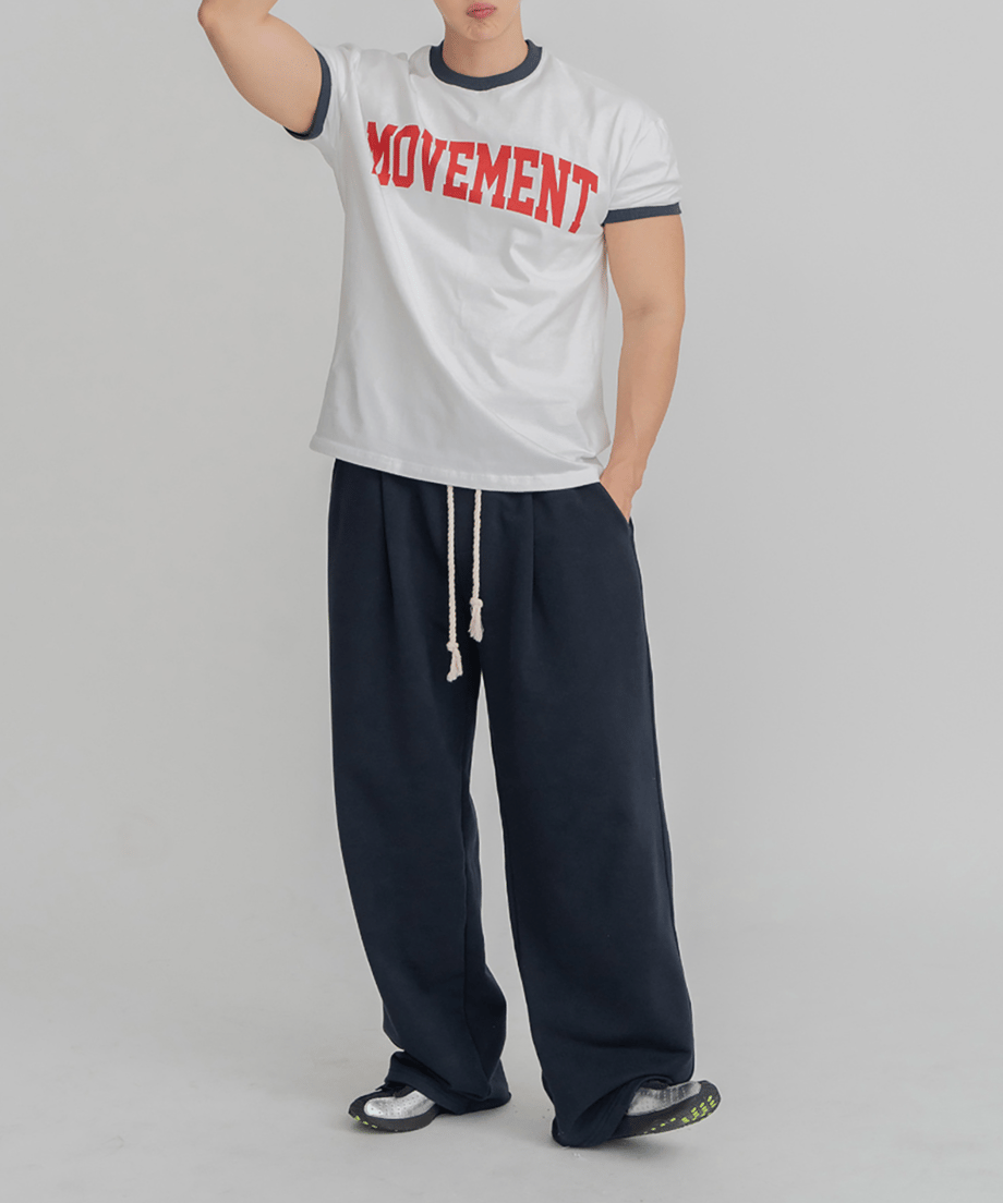 ホワイト地にネイビーのリブ、赤のロゴが入ったリンガーTシャツに、ネイビーのワイドイージーパンツを合わせ、右手を頭に添えて立つ男性の全身スタイリング写真。