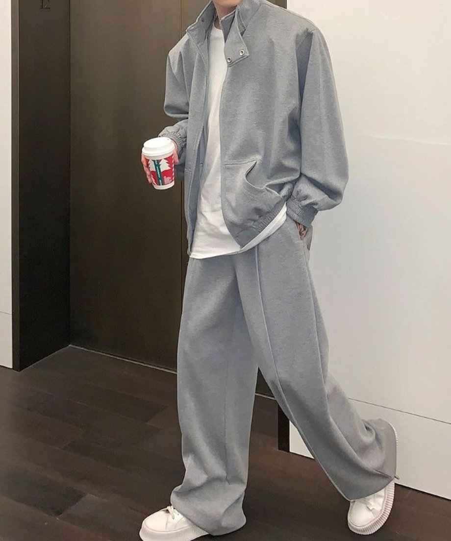 グレーのハイネックジップアップブルゾンと、同色のセンタープレス入りワイドスウェットパンツをセットアップで着用し、白いスニーカーを履いて歩く男性の全身コーディネートカット。