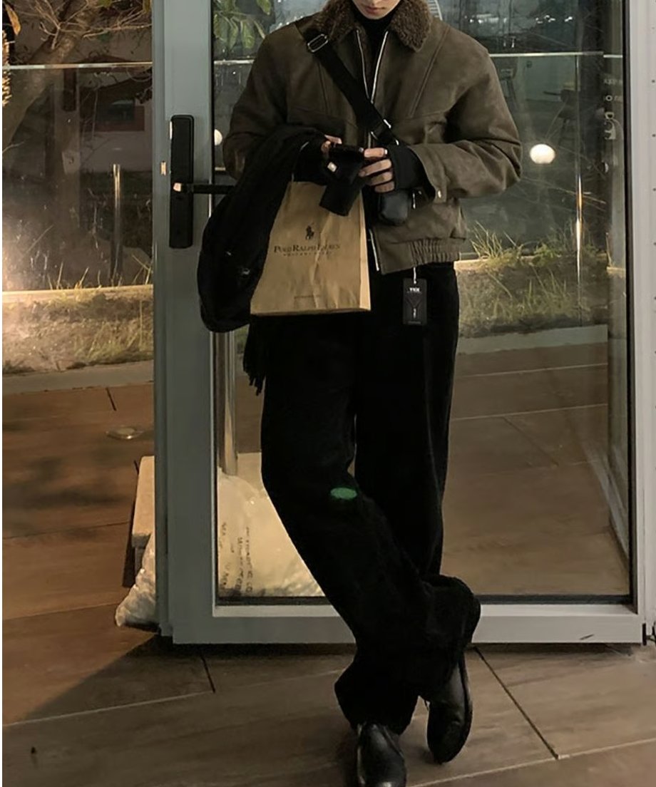 ブラウンのボア襟フライトジャケットに黒のパンツを合わせ、足を交差させてスマートに佇む男性の全身カット。