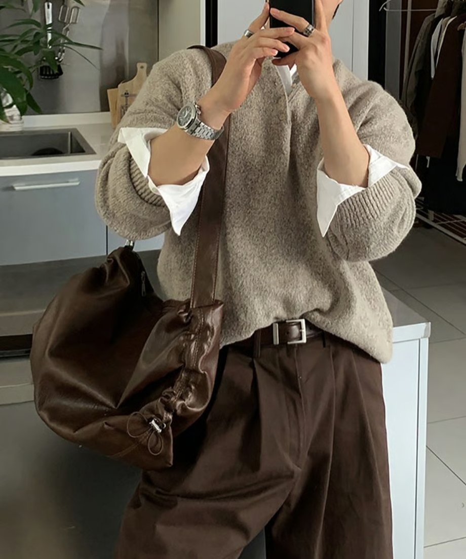室内でベージュのヘンリーネックニットに白シャツ、ブラウンのパンツとベルトを合わせ、スマートフォンで鏡越しに自撮りをする男性のカット。