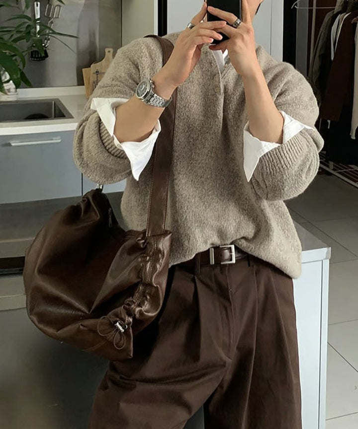室内でベージュのヘンリーネックニットに白シャツ、ブラウンのパンツとベルトを合わせ、スマートフォンで鏡越しに自撮りをする男性のカット。