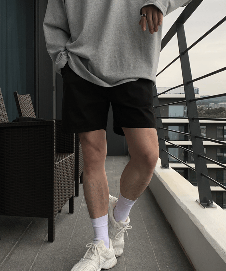 テラスのような場所で、グレーの長袖トップスに黒のショートパンツ、白ソックスと白スニーカーを合わせた男性のスタイリッシュな立ち姿