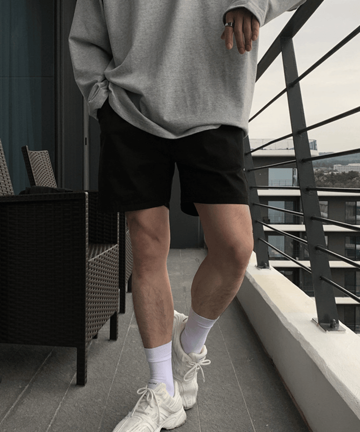 テラスのような場所で、グレーの長袖トップスに黒のショートパンツ、白ソックスと白スニーカーを合わせた男性のスタイリッシュな立ち姿