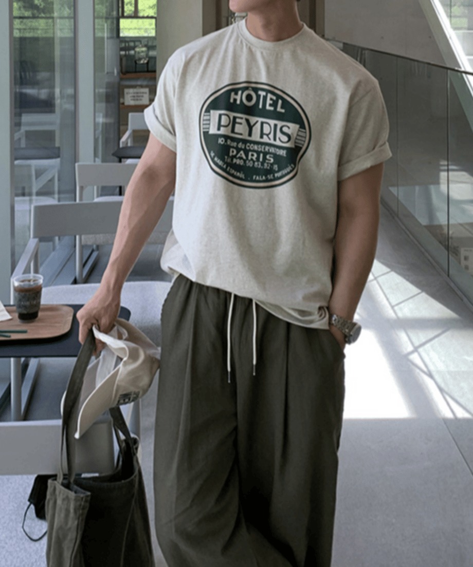 メランジグレーのロゴTシャツの袖をロールアップし、オリーブグリーンのパンツに合わせた男性が、椅子に深く腰掛けて首元に手を添えているリラックスした雰囲気のフロントアップカット。