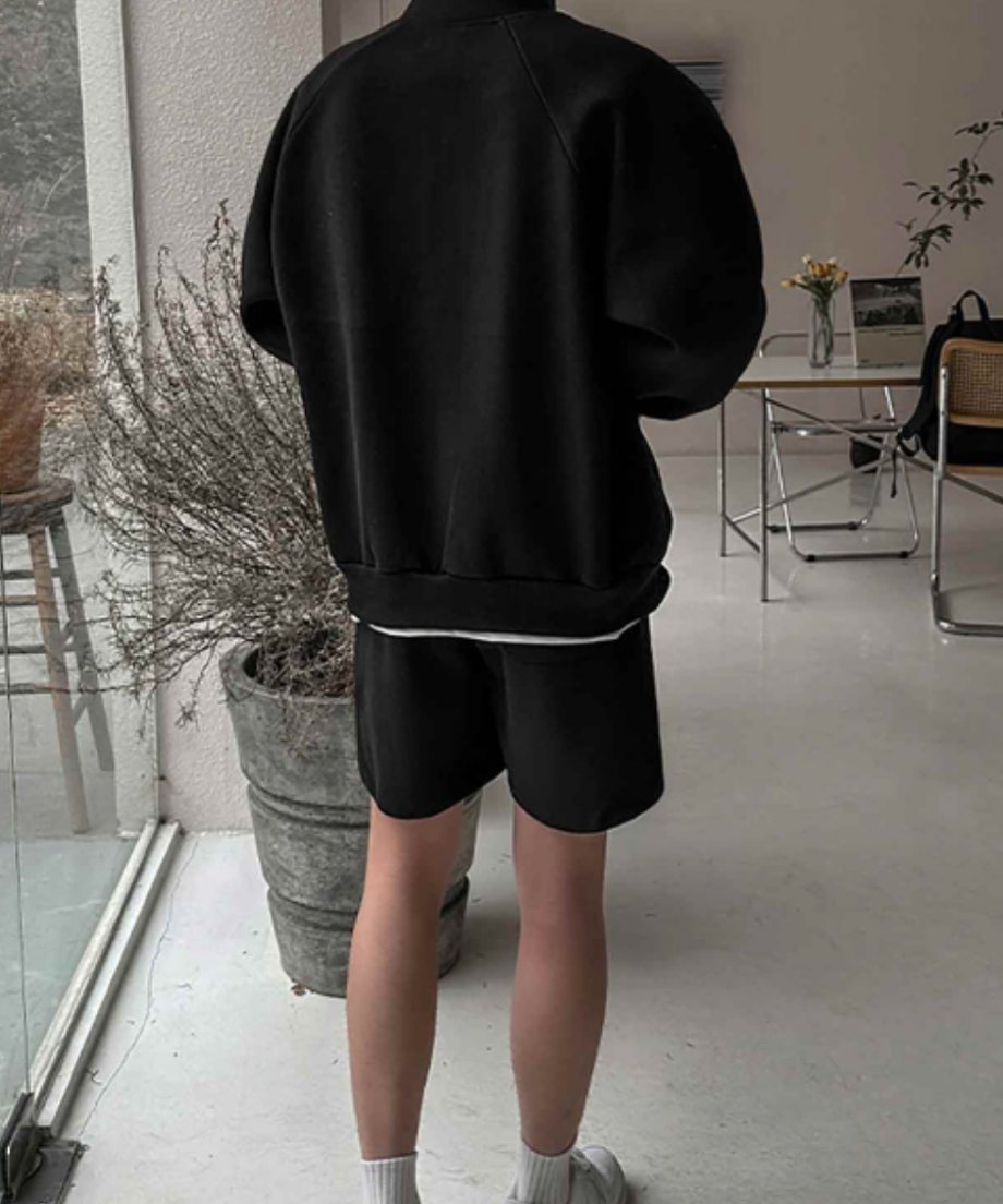 屋内で黒のスタンドカラージップアップジャケットと黒のショートパンツのセットアップを着用した男性のバックスタイル