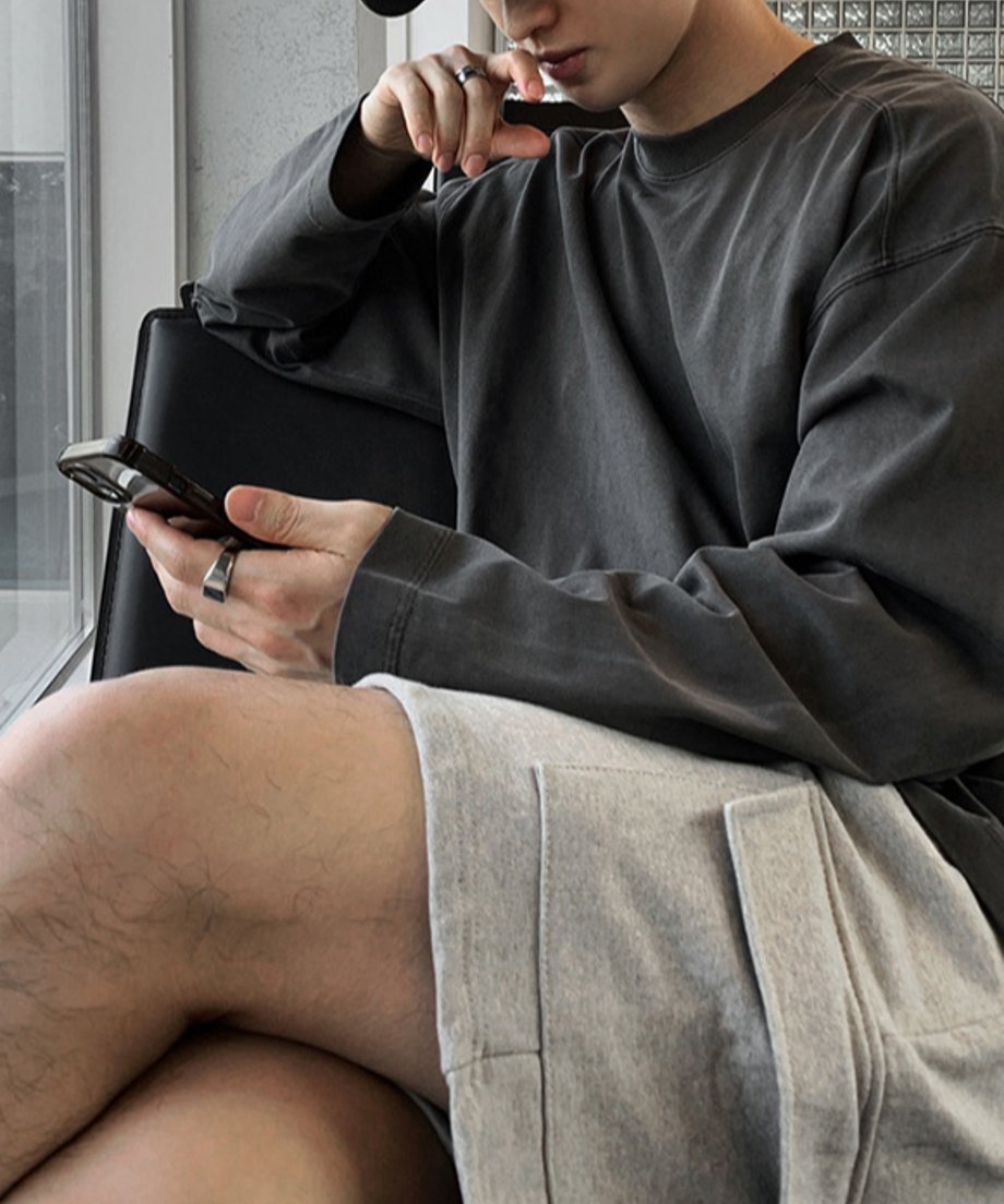 アイアングレーの長袖クルーネックTシャツを着用し、窓際の黒いレザーチェアに座ってスマートフォンを見つめている男性のクローズアップカット。