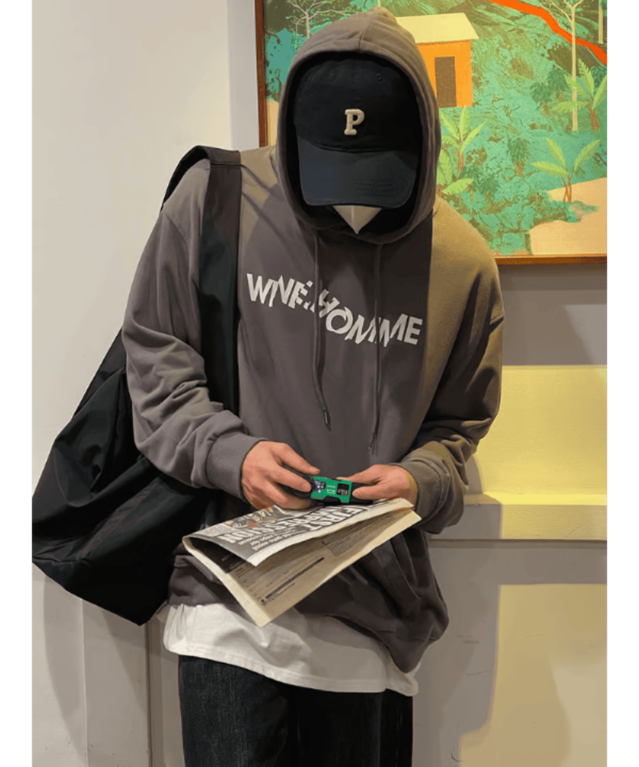 チャコールグレーのロゴ入りプルオーバーパーカーのフードを被り、黒いキャップを合わせた男性が、手元で使い捨てカメラと新聞を持ちながらうつむいている正面チェストアップ。